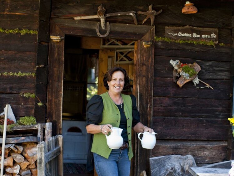 Die Gastgeberin der Hütte steht im rustikalen Holzeingang mit zwei Krügen.