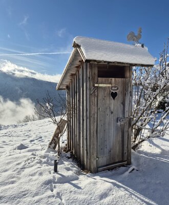 Aussentoilette aus Holz mit verschneitem Dach, Wetterhahn und Herz-Ausschnitt auf dem Hof.
