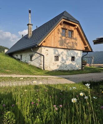 Der Hof mit heller Fassade, Holzgiebel und dunklem Dach, umgeben von einer grünen Wiese.