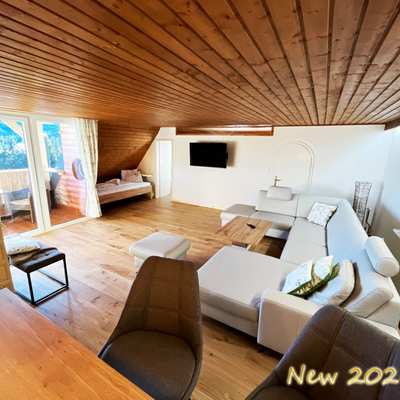 Das Wohnzimmer mit großem Sofa, Fernseher, integriertem Bett, Kachelofen und Balkonzugang.