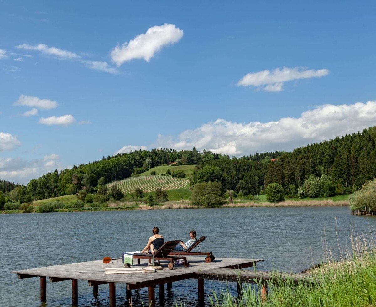 Idyllischer Holzsteg am Teich mit bequemen Liegestühlen und Blick auf die grüne Hügellandschaft.