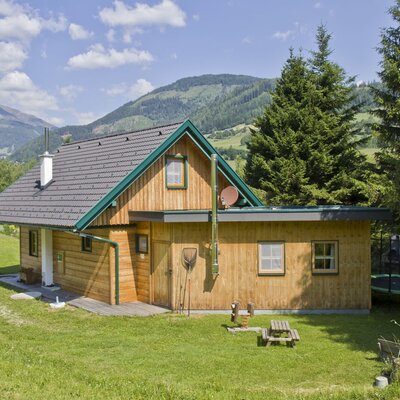 Die Almhütte mit Holzausführung, umgeben von einer Rasenfläche mit Sitzgelegenheiten und einem Trampolin, vor der Bergkulisse.