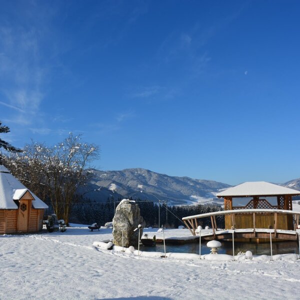 Der verschneite Außenbereich des Bauernhofs mit einer Grillhütte, einem Teich mit Holzpavillon und Blick auf die Berge.