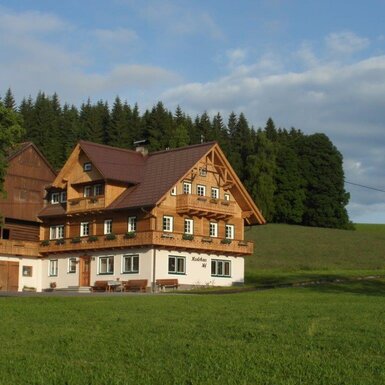 Der Bauernhof mit Holzfassade, Balkonen und Sitzbänken, umgeben von Wiesen und Wald.