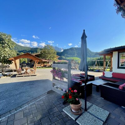 Gepflasterte Terrasse des Bauernhofs mit Outdoor-Sitzgelegenheiten, Sonnenschirm, Bergblick und einem Spielplatz im Hintergrund.