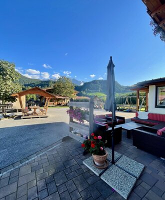 Gepflasterte Terrasse des Bauernhofs mit Outdoor-Sitzgelegenheiten, Sonnenschirm, Bergblick und einem Spielplatz im Hintergrund.