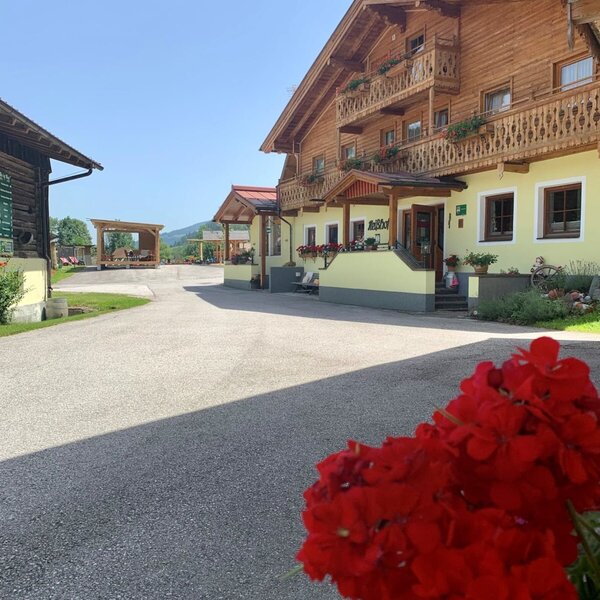 Außenansicht des Bauernhofs mit Holzbalkonen, Zufahrt und einem überdachten Sitzbereich im Hintergrund.