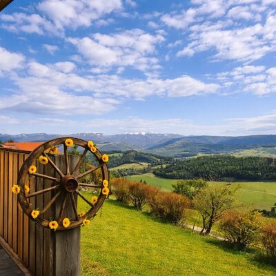 Terrasse des Bed and Breakfast mit Holzzaun, einem dekorativen Wagenrad und Blick auf grüne Täler und ferne Berge.