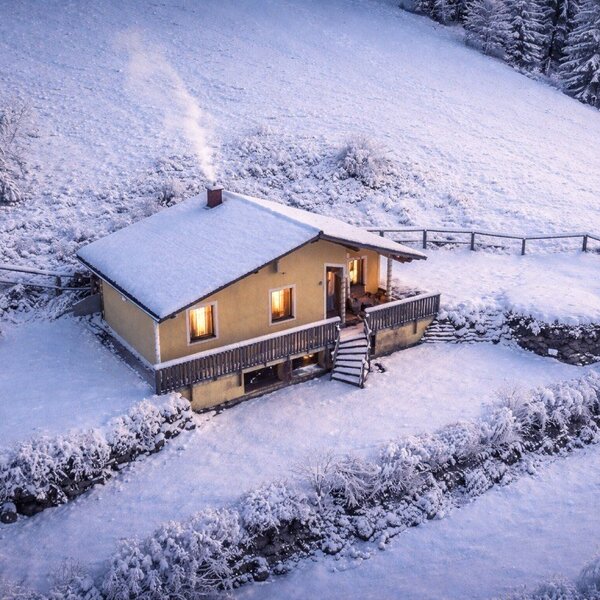 Das Bed and Breakfast im Winter, mit schneebedecktem Dach, rauchendem Schornstein und beleuchteten Fenstern auf einem verschneiten Hügel.