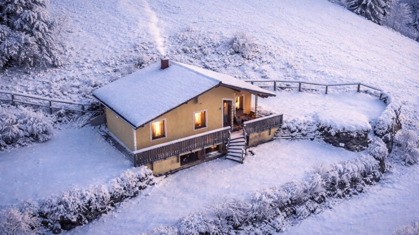 Das Bed and Breakfast im Winter, mit schneebedecktem Dach, rauchendem Schornstein und beleuchteten Fenstern auf einem verschneiten Hügel.