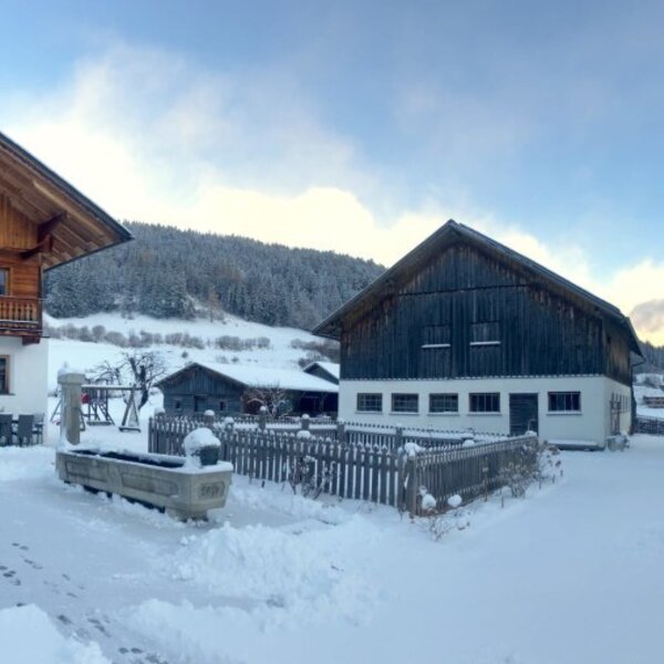 Die schneebedeckte Außenansicht des Bauernhofs mit seinen Gebäuden und der winterlichen Berglandschaft.