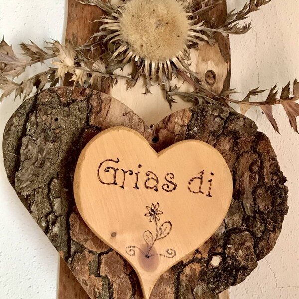 Ein dekoratives Holzherz mit Rinde, der Aufschrift "Grias di" und einer getrockneten Distel im Bauernhof.