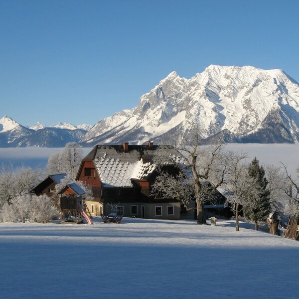 Der Bauernhof in verschneiter Winterlandschaft mit Bergpanorama, Nebeldecke und einem Spielturm.