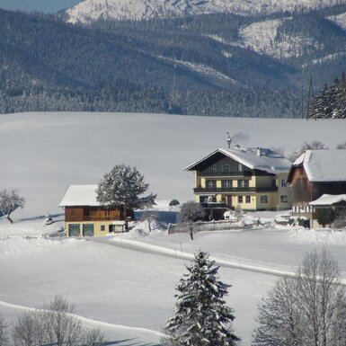 Der Bauernhof in einer weitläufigen, schneebedeckten Winterlandschaft mit Bergen im Hintergrund.