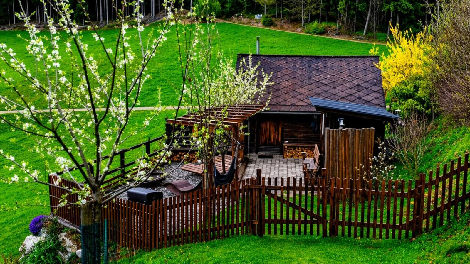 Hütte mit umzäuntem Terrassenbereich, umgeben von grünen Wiesen und Wald.