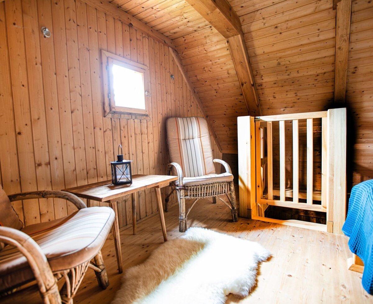 Die Stube der Almhütte ist mit Holzwänden, zwei Korbsesseln, einem Beistelltisch mit Laterne, einem Fenster und einem durch ein Holzgitter abgetrennten Schlafbereich ausgestattet.