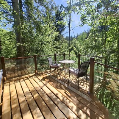 Terrasse mit Holzboden, eine Glastrennwand, einen Tisch und zwei Stühle inmitten der Bäume.