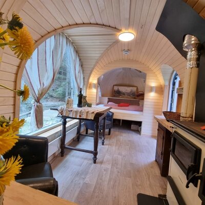 Der Innenraum des Igluhaus mit Holzwänden, einem Essbereich, einem Bett in einer Nische, einem Holzofen und einem großen Fenster mit Waldblick.