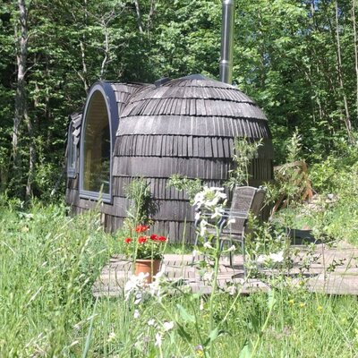 Igluhut mit Holzschindeln, großem Fenster und einer kleinen Terrasse mit Sitzgelegenheiten, umgeben von Natur auf dem Bauernhof.