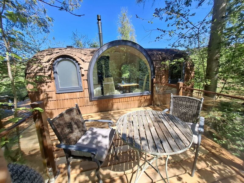 Außenbereich des Igluhuts mit einer Holzterrasse, einem Tisch und Stühlen sowie einer einzigartigen Unterkunft mit großem Panoramafenster inmitten von Bäumen.