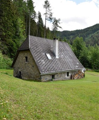 Außenansicht des Hütte mit Steinmauern aus dem Jahr 1800, Schindeldach, Schornstein und einem Stapel Brennholz, umgeben von Wiese und Wald.