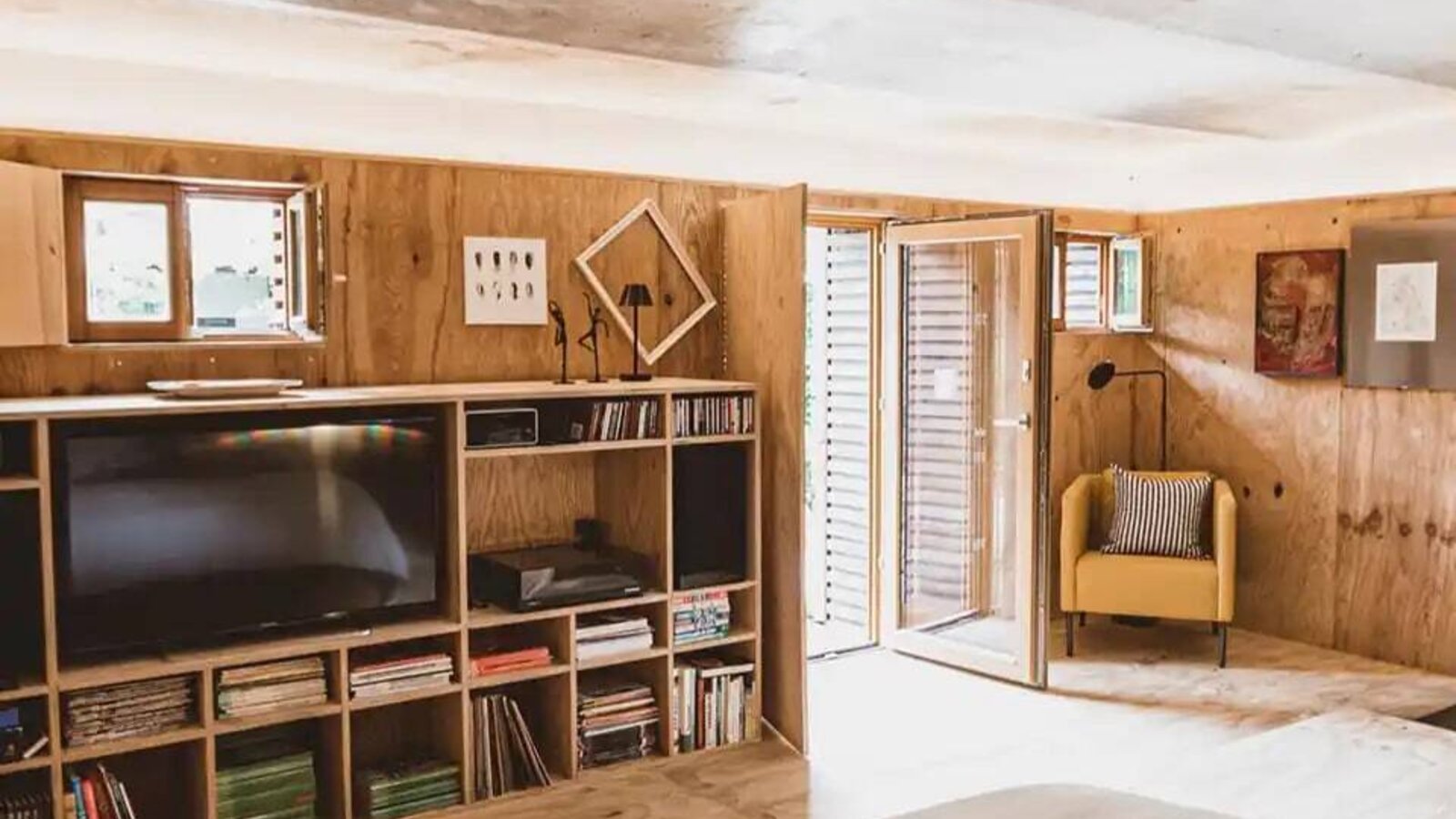 Schlafzimmer im Bauernhof mit großem Bett, Holzwänden, einem Regal mit Fernseher und Büchern, einem gelben Sessel und Zugang nach draußen.