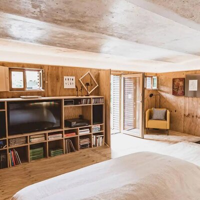 Schlafzimmer im Bauernhof mit großem Bett, Holzwänden, einem Regal mit Fernseher und Büchern, einem gelben Sessel und Zugang nach draußen.