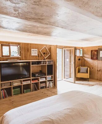 Schlafzimmer im Bauernhof mit großem Bett, Holzwänden, einem Regal mit Fernseher und Büchern, einem gelben Sessel und Zugang nach draußen.