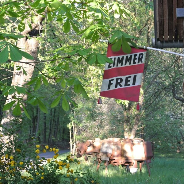 Das 'ZIMMER FREI'-Schild am Bauernhof, umgeben von Bäumen und alten landwirtschaftlichen Geräten.