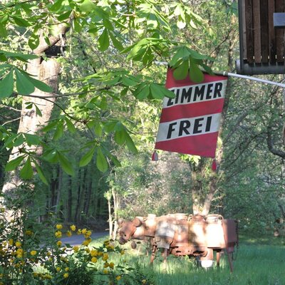 Das 'ZIMMER FREI'-Schild am Bauernhof, umgeben von Bäumen und alten landwirtschaftlichen Geräten.