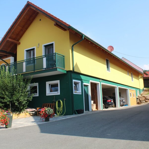Der Bauernhof mit gelb-grüner Fassade, Balkon und Garagenstellplätzen.