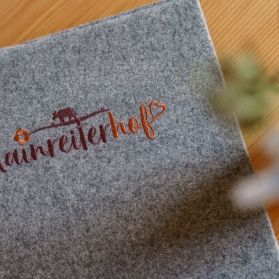 Eine graue Filzmappe mit der Stickerei 'Kainreiterhof' für Gäste auf dem Bauernhof.