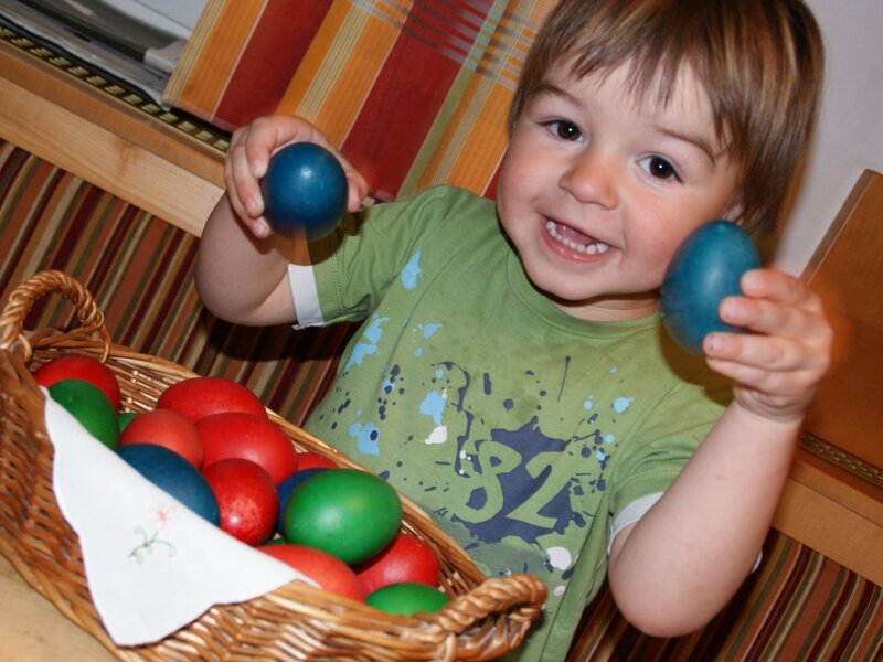 Ein lachendes Kind hält bunte Ostereier neben einem Korb voller Eier auf dem Bauernhof.
