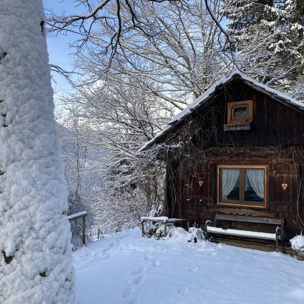 Die verschneite Winterlandschaft umgibt die Holzhütte mit ihren Fensterbalken, dem roten Herz und der gemütlichen Bank davor.