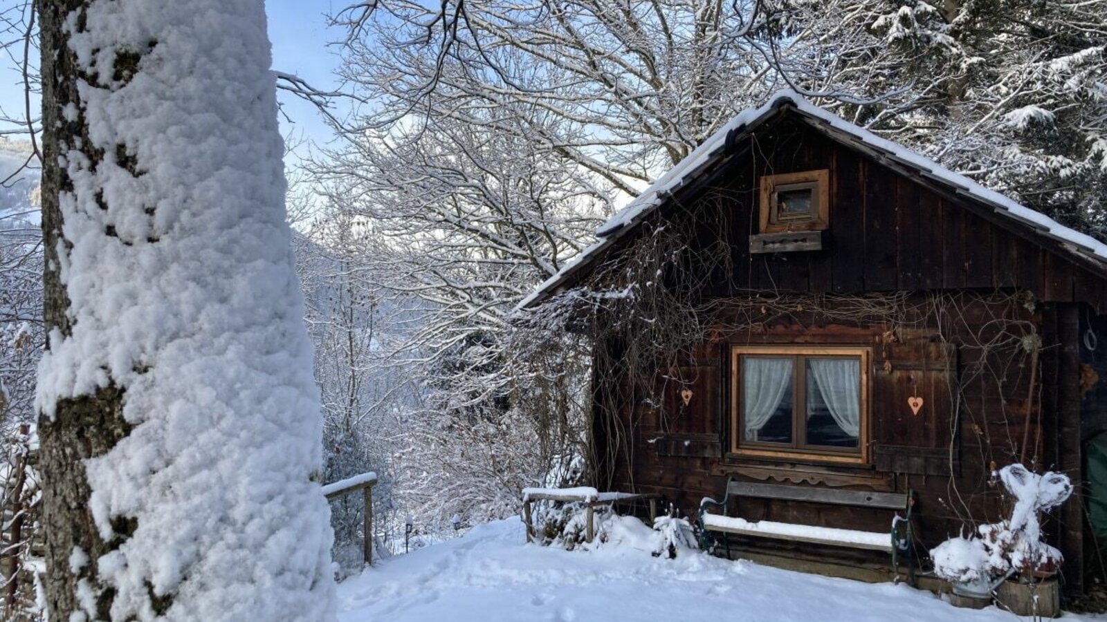 Die verschneite Winterlandschaft umgibt die Holzhütte mit ihren Fensterbalken, dem roten Herz und der gemütlichen Bank davor.