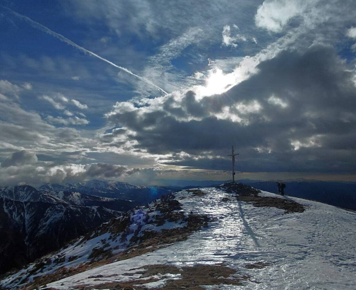 Blick auf einen verschneiten Berggipfel mit Gipfelkreuz_Greim, 2474m_und weitem Bergpanorama unter einem bewölkten Himmel.