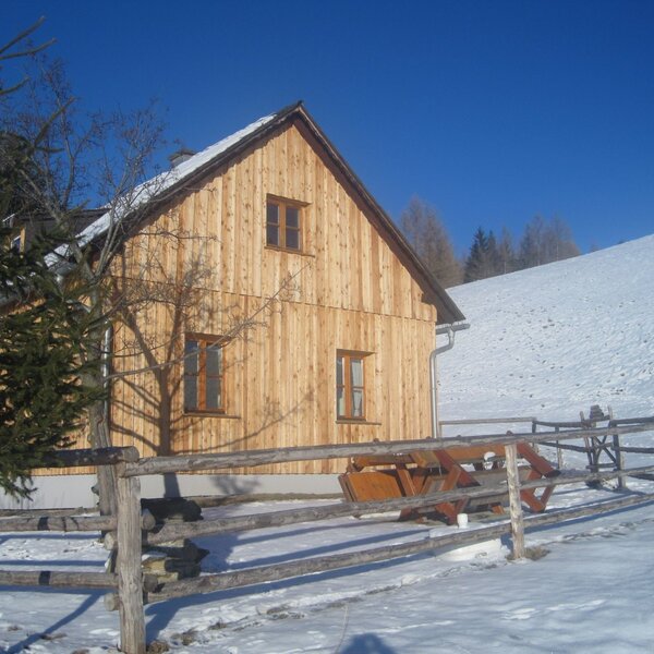 Die hölzerne Almhütte im Winter, mit einer Sitzgruppe im umzäunten Außenbereich und schneebedeckten Hängen.