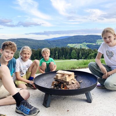 Kinder sitzen um eine Feuerstelle auf dem Bauernhof mit Blick auf die Berge.