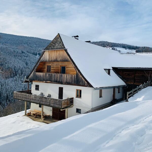 Außenansicht des schneebedeckten Bauernhauses mit Holzbalkon und Ausblick auf die bewaldeten Hügel.