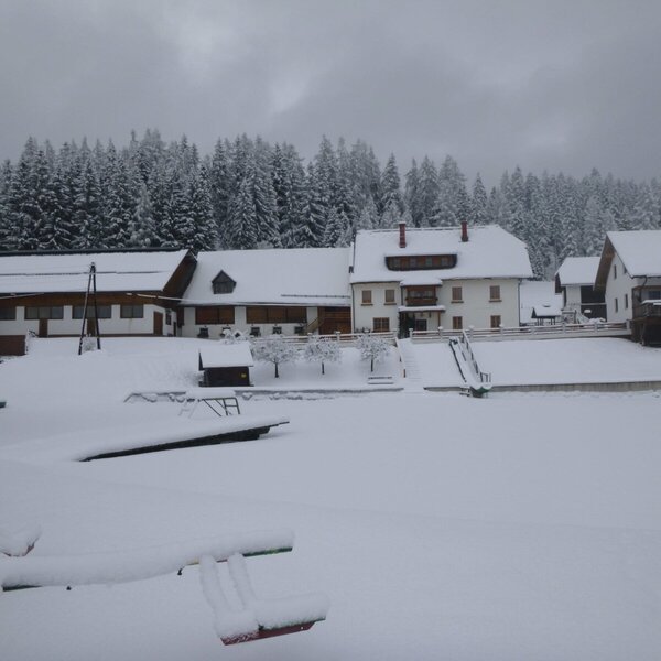 Der verschneite Bauernhof im Winter, umgeben von Wald und einem zugefrorenen Gewässer mit Booten im Vordergrund.