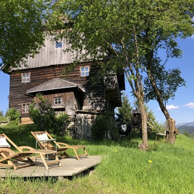 Außenansicht mit einer Holzterrasse, zwei Liegestühlen und einem Tisch, umgeben von grüner Wiese und Bäumen, mit Bergblick.