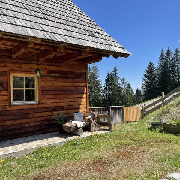 Außenansicht der Alm mit Holzfassade, Fenster, Holzstapel und einer Sitzbank aus Holzstämmen.