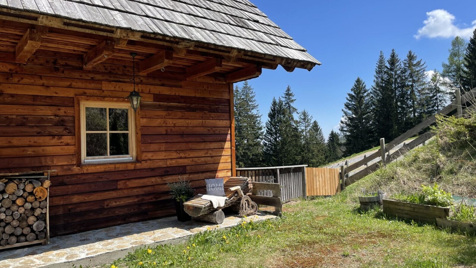 Außenansicht der Alm mit Holzfassade, Fenster, Holzstapel und einer Sitzbank aus Holzstämmen.