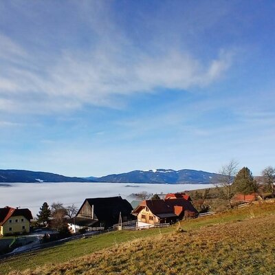 Die Umgebung des Bauernhofs mit Dorf, Kirche, Nebel im Tal und Bergen im Hintergrund.
