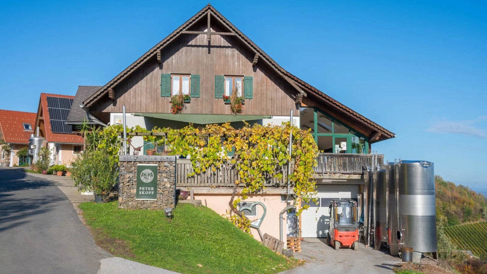 Außenansicht des Bauernhofs mit Holzfassade, Weinstöcken und Weintanks, die auf die Weinproduktion des Weinguts hinweisen.