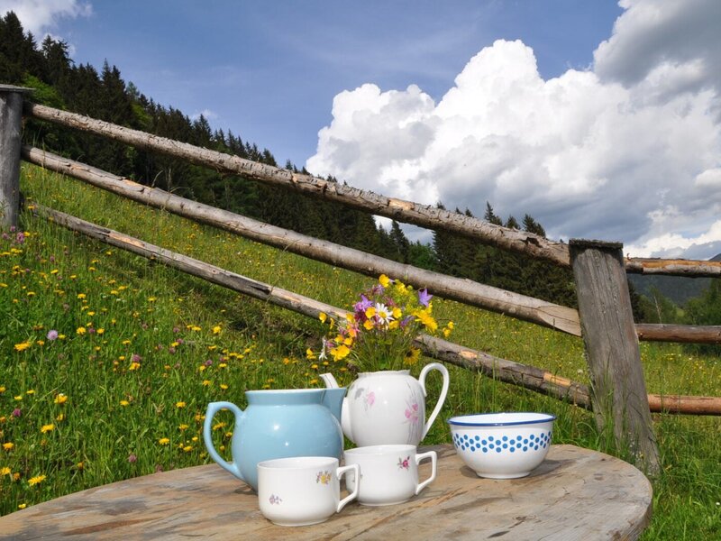 Tisch im Freien mit Geschirr und Blumenstrauß vor der Kulisse einer blühenden Wiese und einem Holzzaun am Hof.