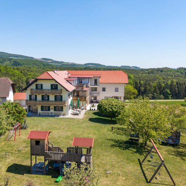 Der Bauernhof mit Balkonen und Außenterrasse, umgeben von einem Garten mit Kinderspielplatz und der Landschaft des Pöllauer Tals.