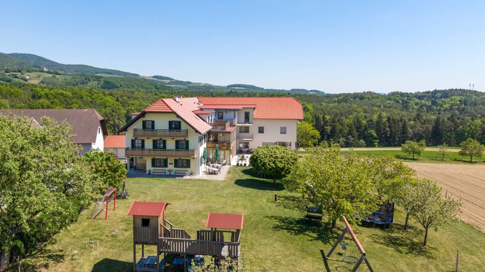 Der Bauernhof mit Balkonen und Außenterrasse, umgeben von einem Garten mit Kinderspielplatz und der Landschaft des Pöllauer Tals.