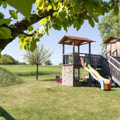 Kinderspielplatz auf dem Bauernhof mit einem hölzernen Spielturm, einer bunten Rutsche und einer Kletterwand, umgeben von grünen Feldern.