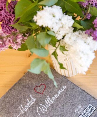 Filzmatte mit der Aufschrift 'Herzlich Willkommen' und dem 'Urlaub am Bauernhof' Logo, begleitet von einem Fliederstrauß auf einem Holztisch.